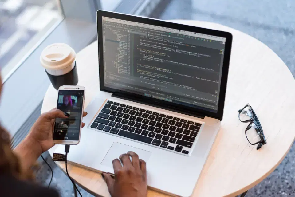 Developer holding a phone while Python code runs on a laptop at a cafe table