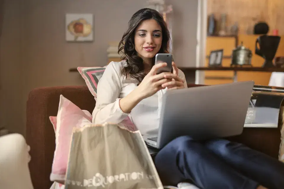 Woman shopping on her phone with a laptop and shopping bag, like a Shopify store customer