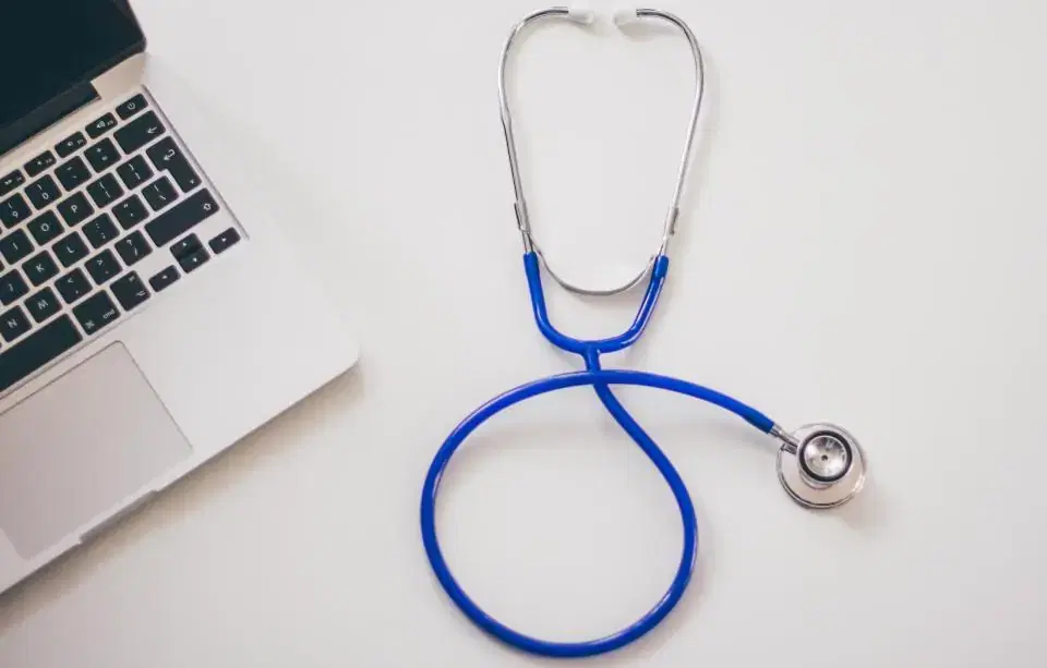 Blue stethoscope lying next to a laptop on a white desk, representing healthcare software
