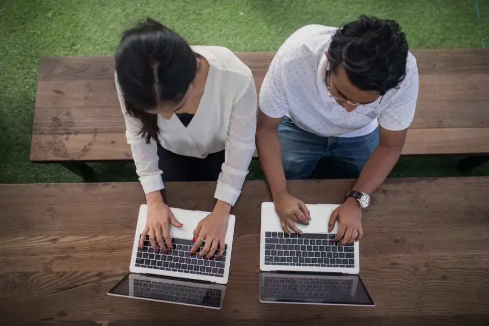 Two developers working side by side on laptops at a wooden table, building AI software