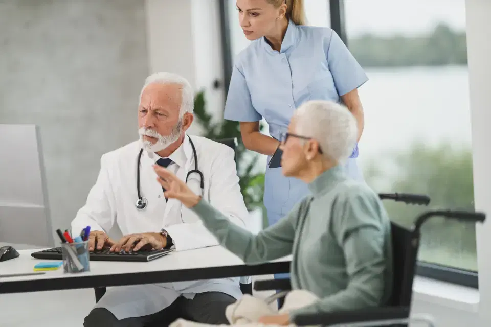 Doctor at a computer consulting an elderly patient while a nurse assists in a clinic