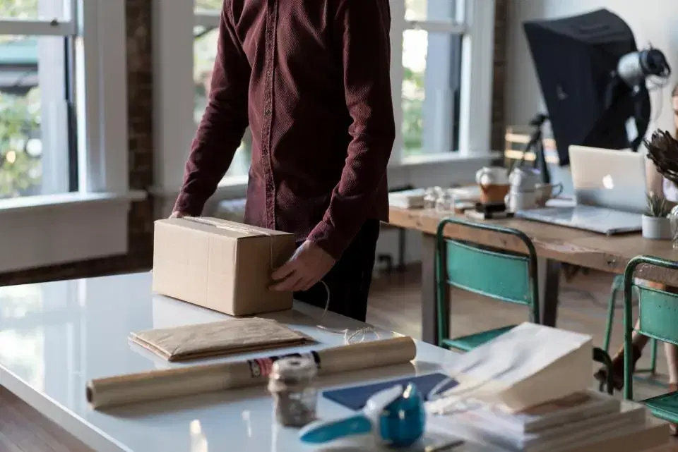 Person packing an online order into a cardboard box on a desk beside a laptop