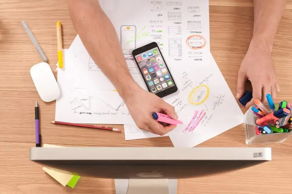 Designer sketching mobile app wireframes on paper beside a smartphone and desktop computer
